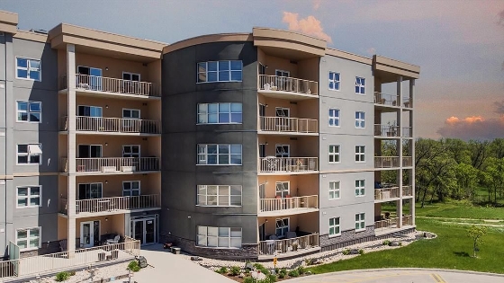 The exterior of 130 Creek Bend Road in River Park South, Winnipeg. The condominium is 5 floors high, and follows a pattern of a light grey wall with two small windows, a column of balconies, and a dark grey wall with one large horizontal window. The property is nicely landscaped. 