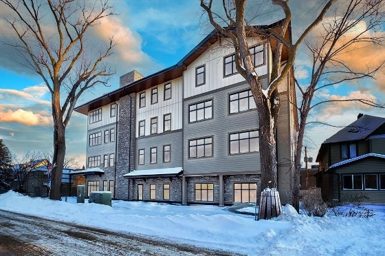 Exterior of 227 Stafford Ave in Crescentwood, Winnipeg. There is snow on the ground, slightly frosted trees and a sunny sky. The condominium is a mix of light and dark grey with brick accent. 