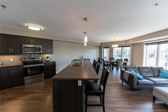 The interior of 227 25 Bridgeland Drive North in Bridgwater Forest, Winnipeg. Open concept with the kitchen and island, living room, and dinning area in view. Dark wooden flooring throughout, dark cabinetry in the kitchen, and an earthy colour scheme throughout the condo. There are floor to ceiling windows in the dinning area and living room.