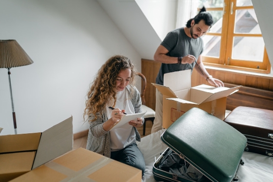 A couple packing their bedroom. The woman is checking off item on her inventory list. The man is filling a box.