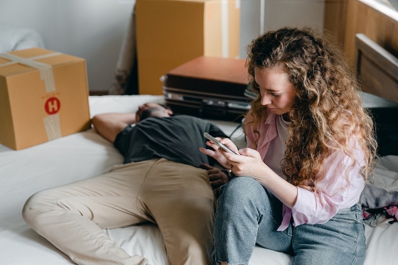 A couple on their bed surrounded by moving boxes. The man is laying down. The woman is sitting beside him on her phone.