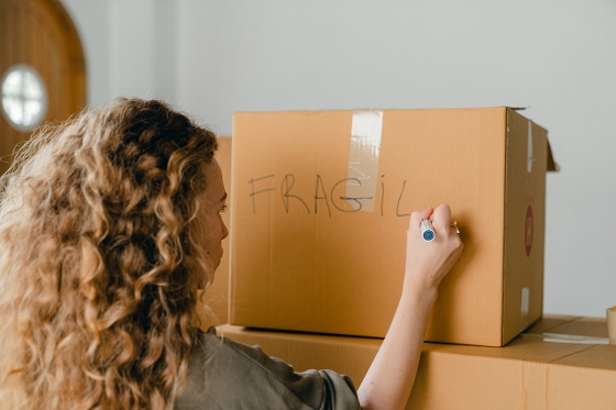 A woman with curly hair writing "Fragile" on a cardboard box in black marker.