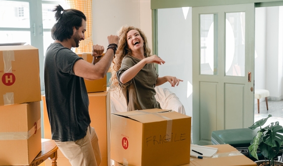 A couple packing boxes. The are looking at each other and laughing. Boxes surround them, the one in front of them says "fragile" in black marker.