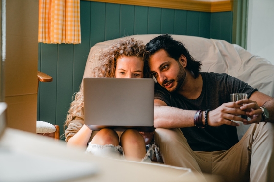 A couple looking at a lap top together while sitting on the floor. There is a dark green wall behind them. They're sitting on a white bean bag chair.