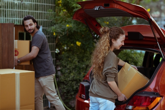 A couple packing their red car with moving boxes. The man is grabbing boxes near a fence, while the woman puts boxes in the trunk.