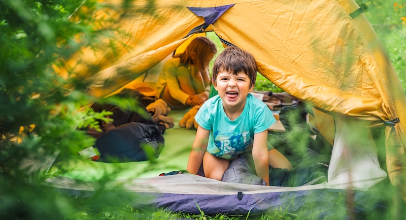 A little boy sticks his head outside of a yellow tent, with his tongue sticking out. There is a young girl in the tent behind him as well.