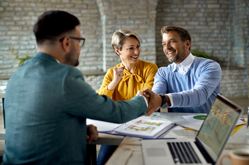 A couple and realtor sit at a table. The husband is shaking hands with said realtor, across the desk. They are smiling.