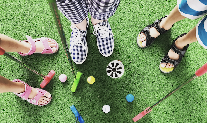 A top-down shot of two kid's feet in sandals and shoes. They are holding mini golf putters and there are colourful golf balls.