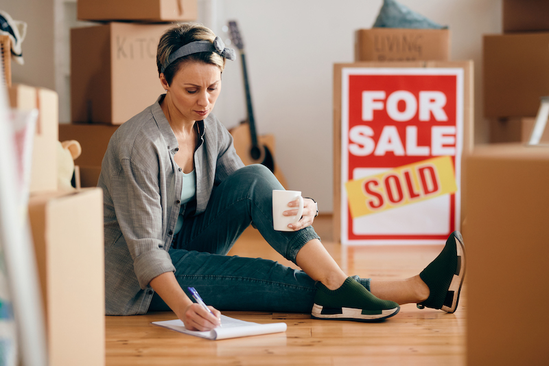 A woman sits on the floor of her home, with a coffee in one hand and writing a list with the other. There are packing boxes all around her and a red "For Sale - Sold" sign.