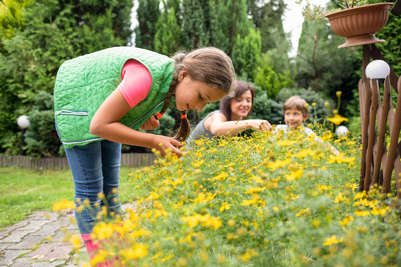 A little girl in a green vest bends down to smell the yellow flowers. Her mom and brother are in the background.